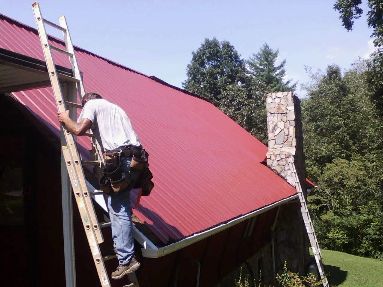 Roofer on a ladder beside a red metal roof and stone chimney — Soffit & Fascia Repair by Roofing Brothers in Sykesville